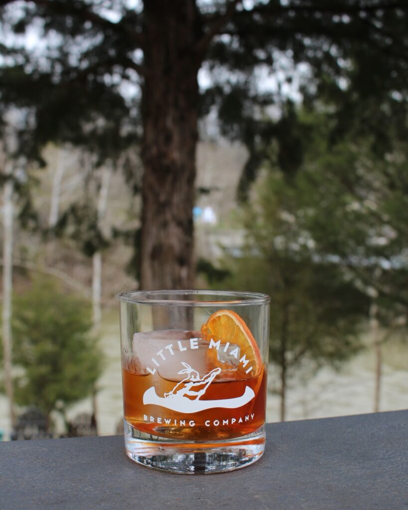 A smiling man holding his dog by the river with two glasses of Little Miami Brewing Company’s beer on the table.