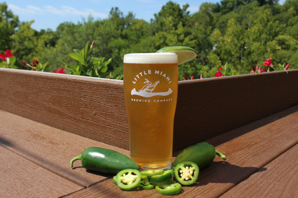 A glass of Piñata jalapeño beer surrounded by fresh whole and sliced jalapeños on a wooden patio table at Little Miami Brewing Company.