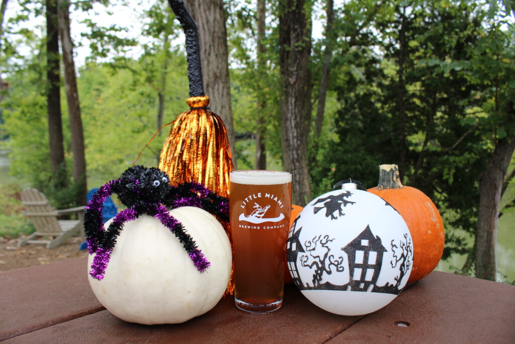 A glass of LMBC’s Witches Brew Java Pumpkin Ale on a rustic wooden table, surrounded by Halloween props like skulls and cobwebs.