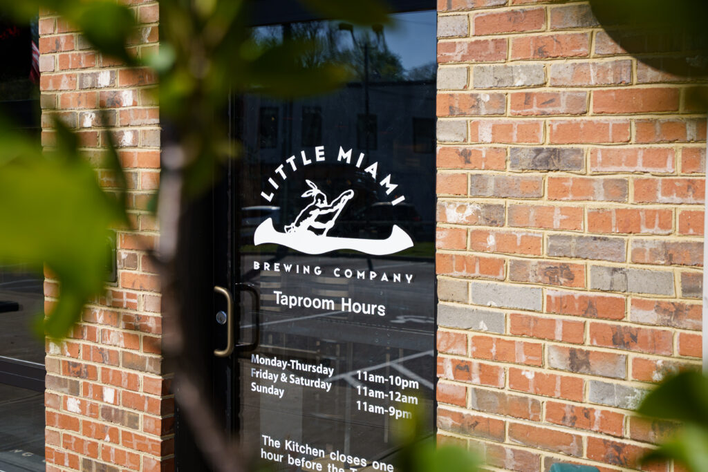 Glass entrance door of Little Miami Brewing Company with canoe-themed logo and printed taproom hours.