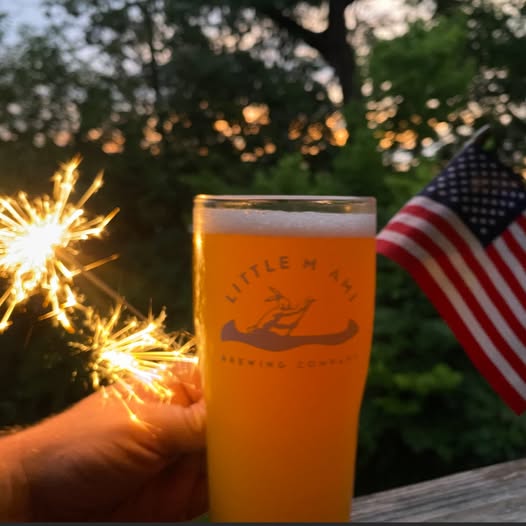 Glass of Little Miami Brewing beer with fireworks, an American flag, and sunset skies in the background during a Fourth of July celebration.
