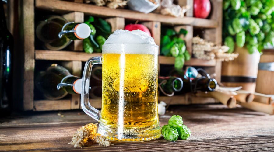 A frothy mug of golden beer sits on a rustic wooden surface, with hops and barley nearby. Bottles in a wooden rack add to the setting, subtly hinting at the natural ingredients that can influence diacetyl flavors in beer.