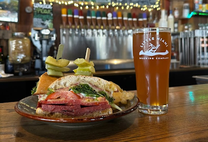 Sandwich with tomato, lettuce, and meat beside a beer glass from Little Miami Brewing Co. Menu board in background.