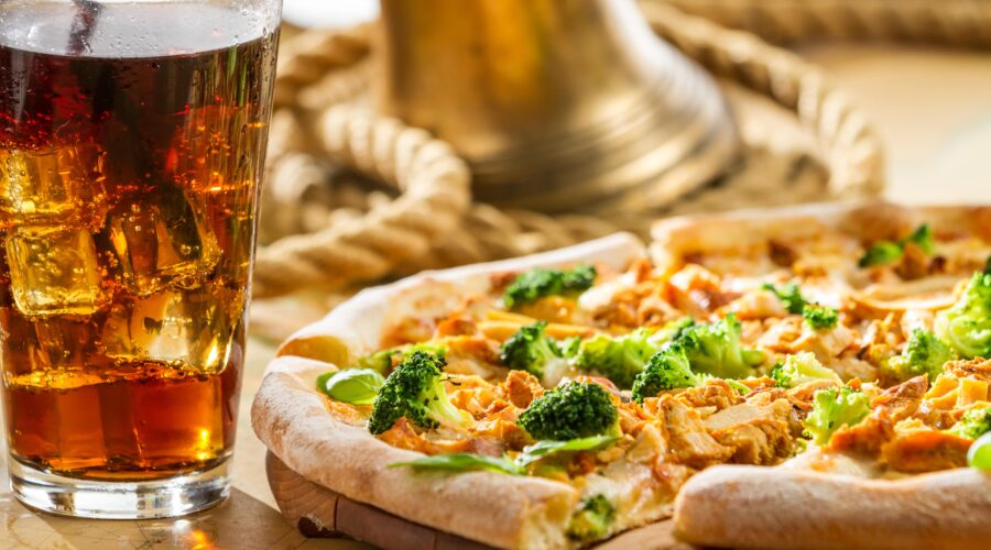 A freshly poured glass of beer with a foamy head sits alongside a hot pizza topped with broccoli and chicken. This pairing combines the richness of beer with the savory flavors of pizza, enhancing the meal experience.