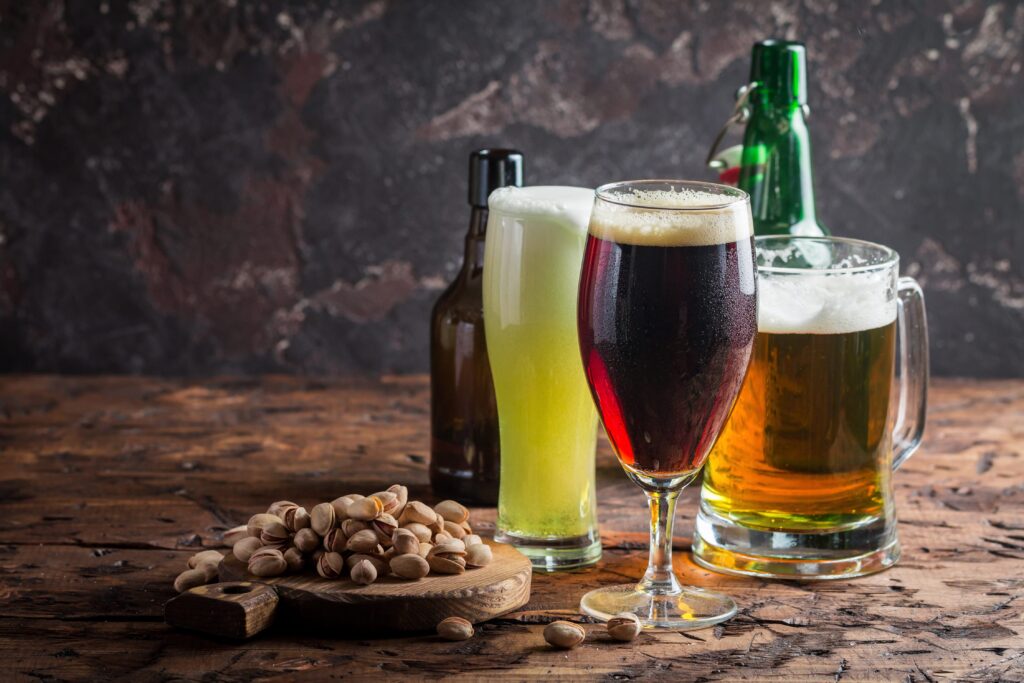 Three different beers are displayed on a wooden table. There's a light beer in a bottle, a dark beer in a glass, and a light beer in a mug. A bowl of pistachios sits in the foreground.