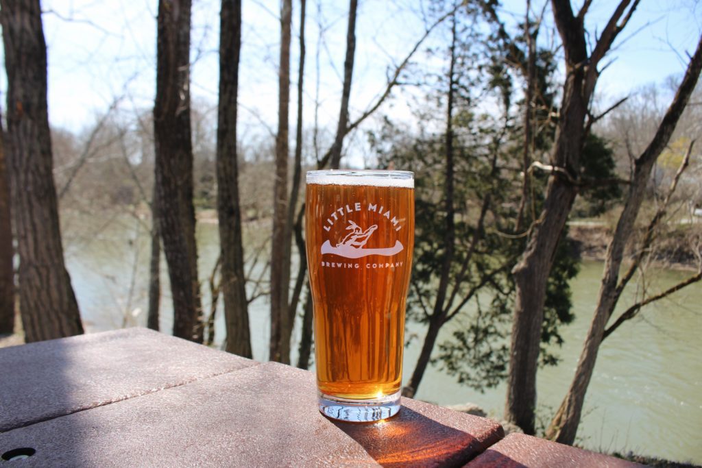 An amber colored beer inside a Little Miami Brewing Company Glass in front of a scenic river backdrop