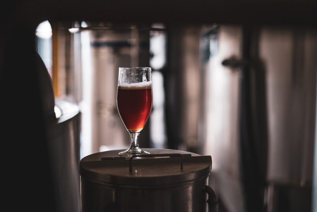 A dark brown beer in a glass, on top of a brewing canister