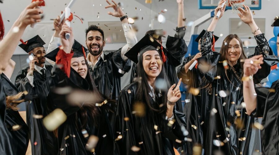 A group of diverse, joyful graduates in black caps and gowns are celebrating their graduation indoors. Confetti fills the air as they cheer, raise their diplomas, and smile widely. The festive atmosphere captures the excitement and happiness of completing their studies, symbolizing a lively "celebrate your graduation" moment.