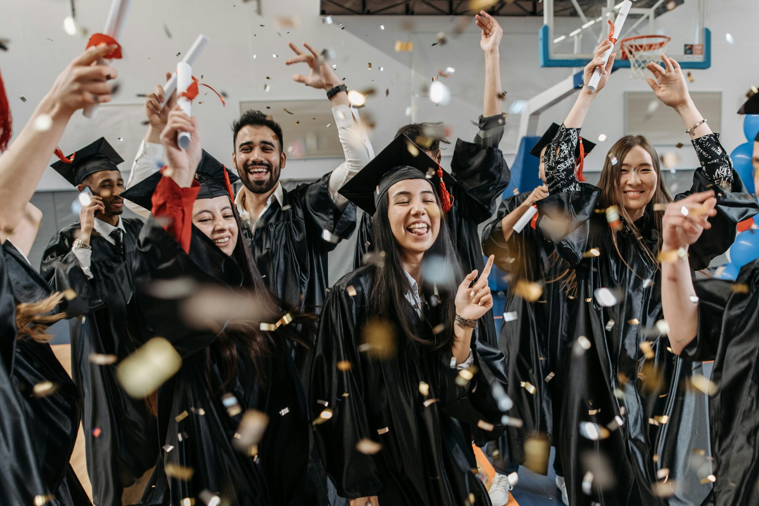 A group of diverse, joyful graduates in black caps and gowns are celebrating their graduation indoors. Confetti fills the air as they cheer, raise their diplomas, and smile widely. The festive atmosphere captures the excitement and happiness of completing their studies, symbolizing a lively "celebrate your graduation" moment.