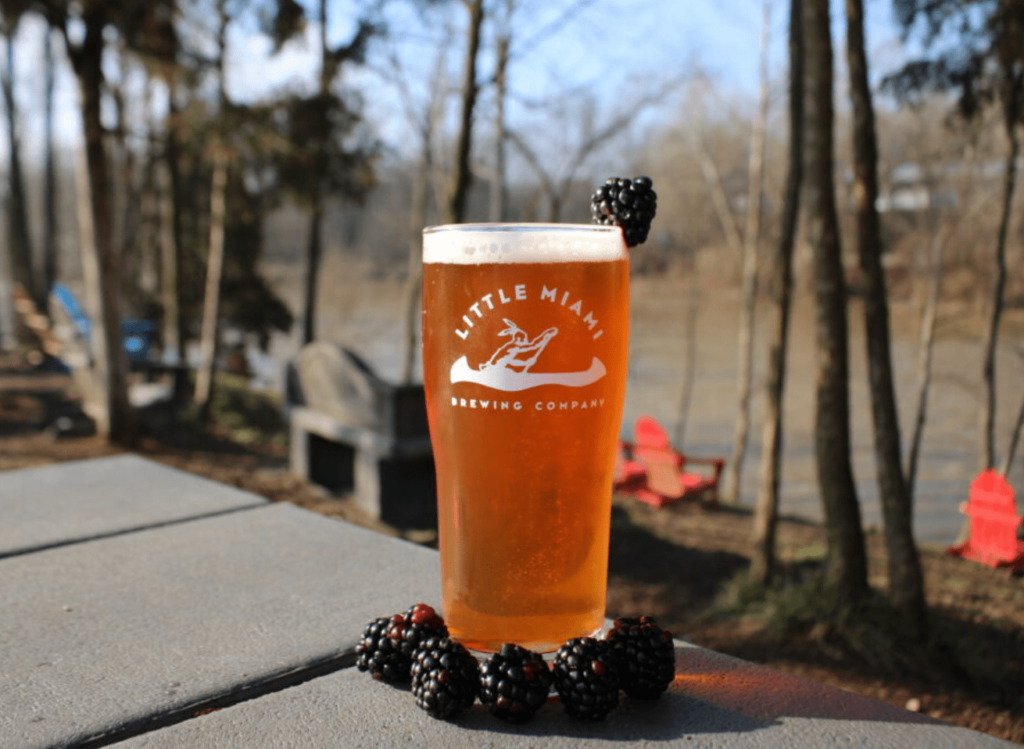 A pint of craft beer from Little Miami Brewing Company, Milford, Ohio, garnished with blackberries on a patio overlooking a scenic riverside with trees and Adirondack chairs in the background.