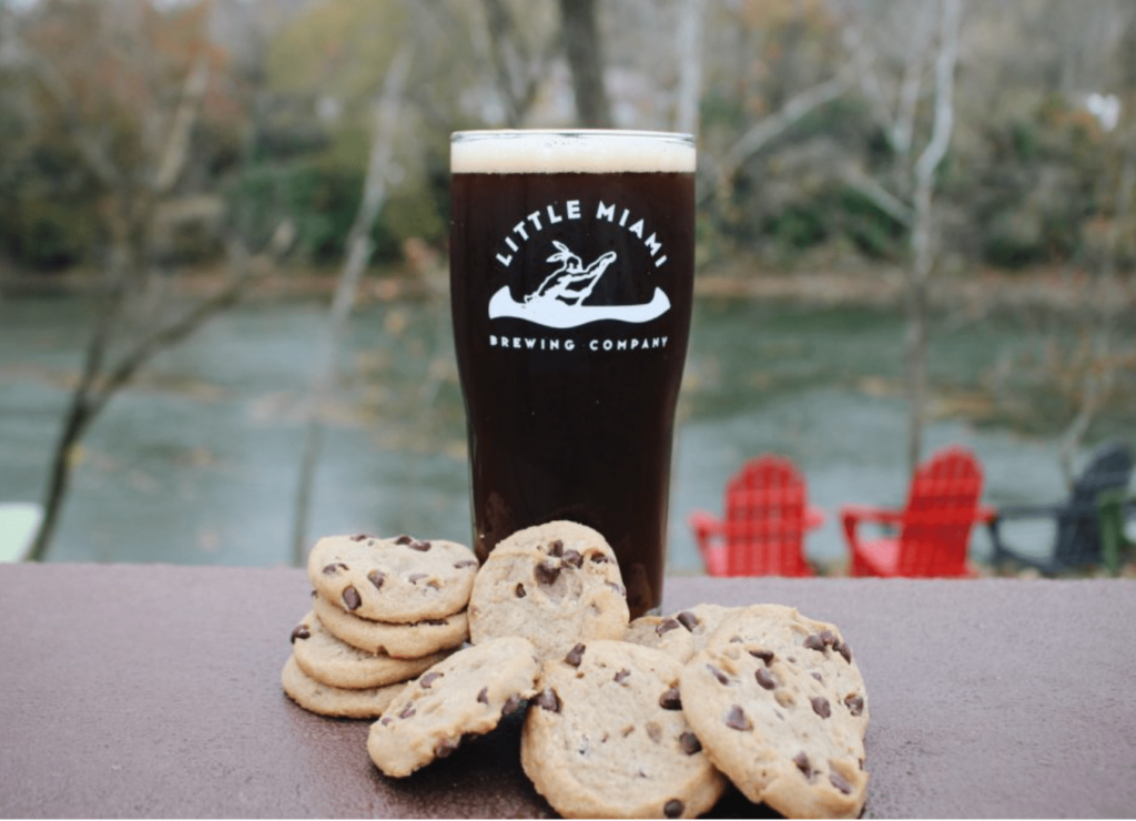 A glass of dark craft beer from Little Miami Brewing Company paired with chocolate chip cookies, set against a riverside backdrop with red Adirondack chairs and trees. Perfect for brewery trips near me.






