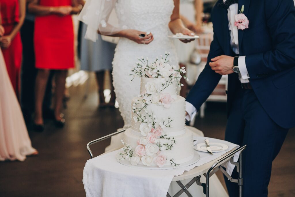 A three-tiered wedding cake decorated with pink and white roses, topped with baby's breath, as the bride and groom prepare to serve.