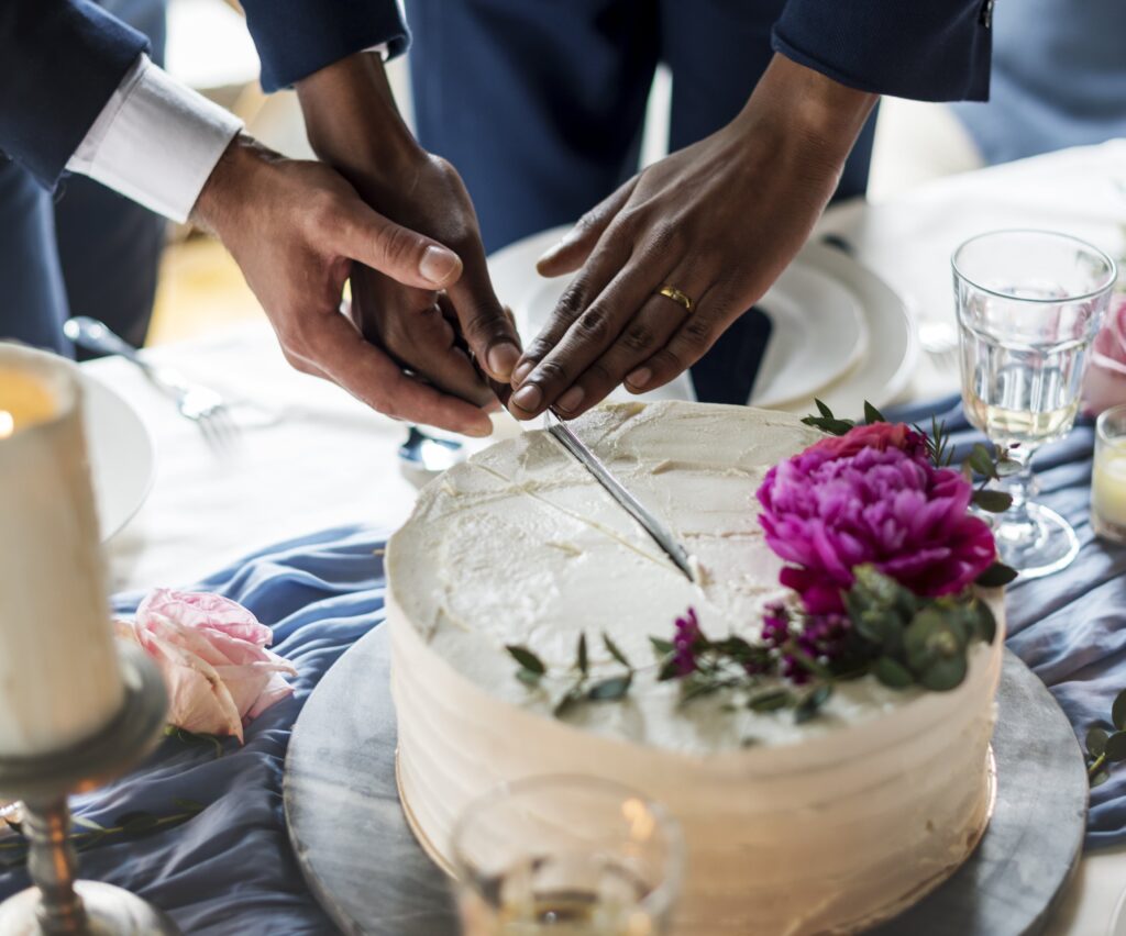 Bride and groom cutting a simple wedding cake topped with vibrant pink flowers. A sweet moment captured in their celebration.