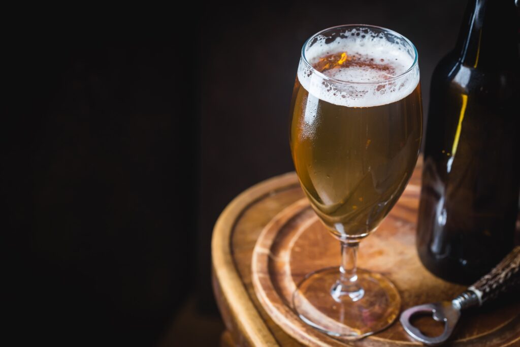 A glass of beer with foam on top, a bottle, and a bottle opener on a wooden board.