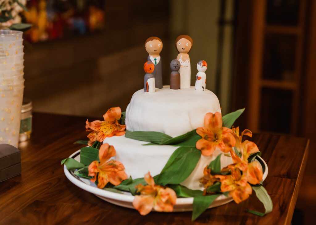 A two-tiered wedding cake decorated with orange flowers and greenery. The top tier features wooden figurines representing the bride, groom, and their dog.