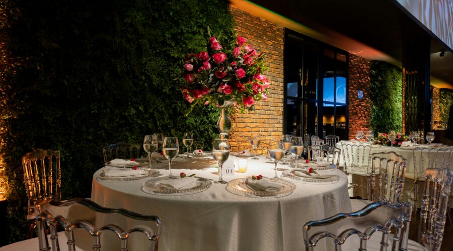 A beautifully set dinner table with a tall floral centerpiece, surrounded by chairs and a green wall.