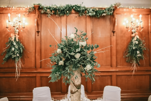 A large floral centerpiece with white roses and greenery sits on a wooden table in a reception venue. The venue has wood-paneled walls and elegant chandeliers.