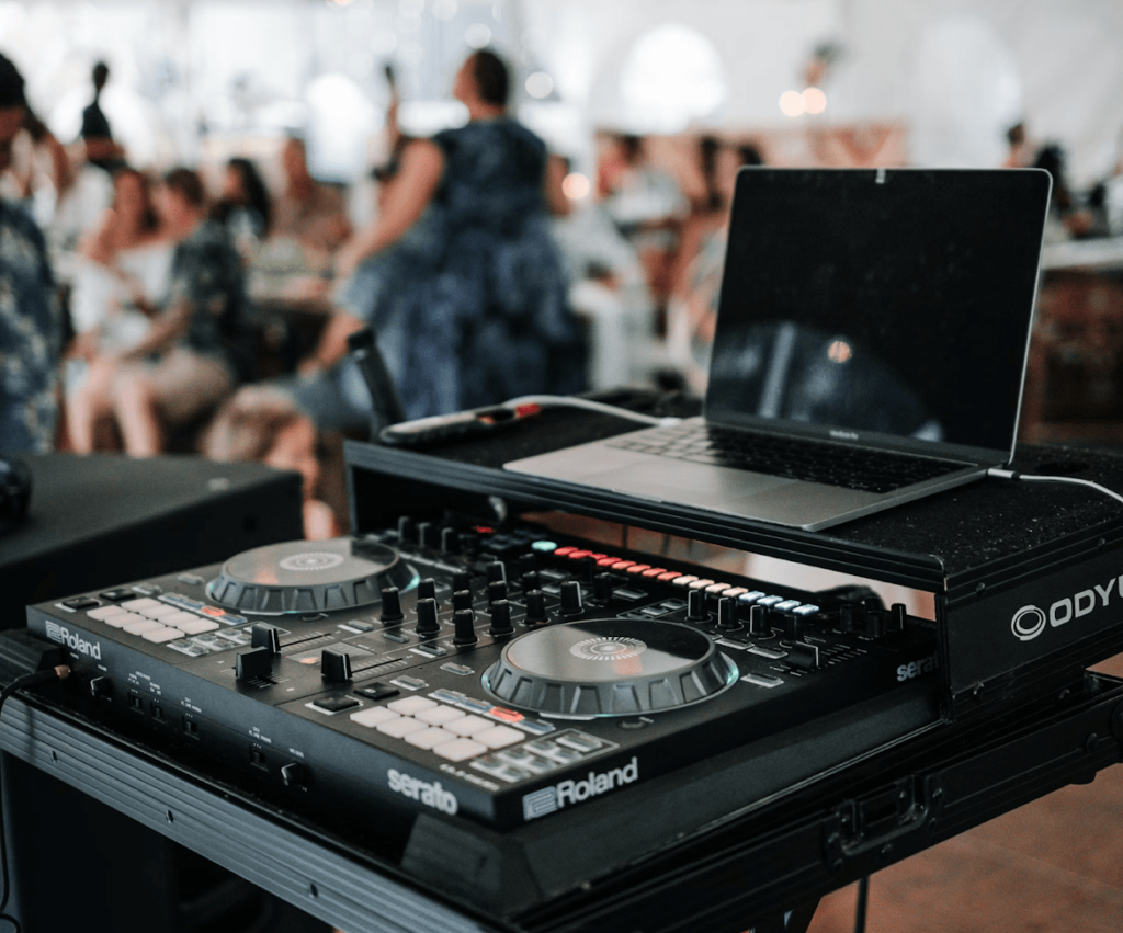 DJ setup at a lively reception venue, featuring a laptop and a Roland Serato controller, with blurred guests mingling in the background.