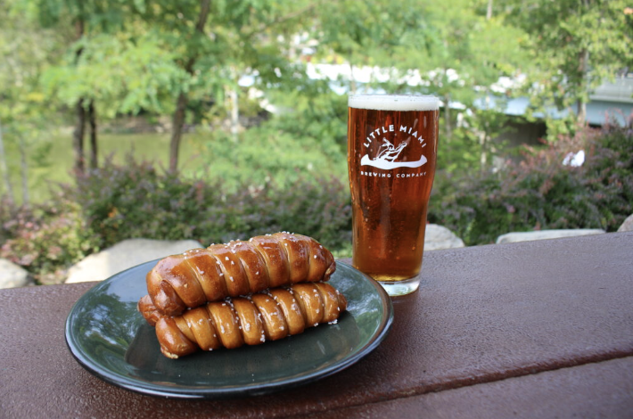 A glass of beer from Little Miami Brewing Company paired with a plate of pretzels on an outdoor table with a scenic background.