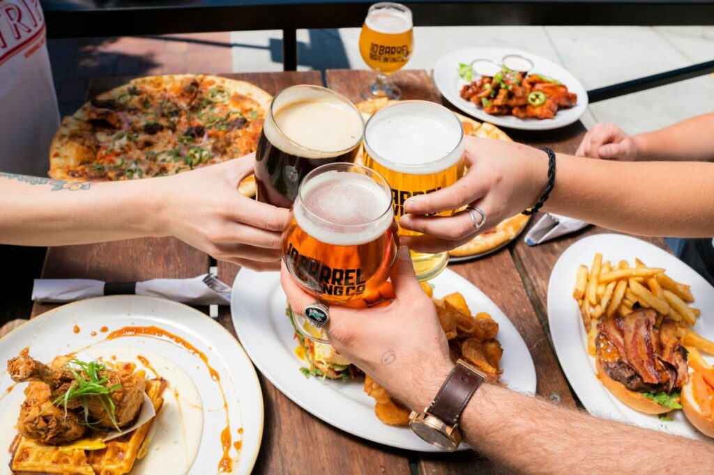 People toasting with glasses of beer at a table with food, including pizza, wings, burgers, and fries.