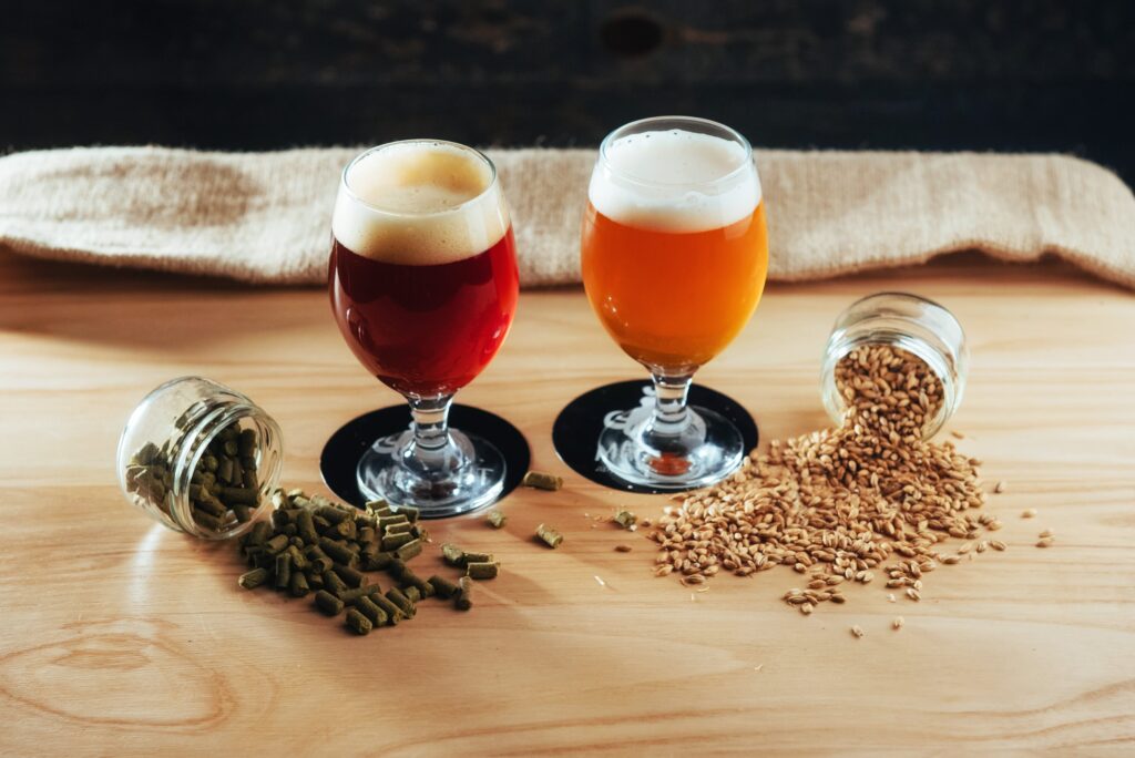 Two glasses of seasonal brews, one amber and one golden, paired with spilled jars of hops and barley on a wooden table.