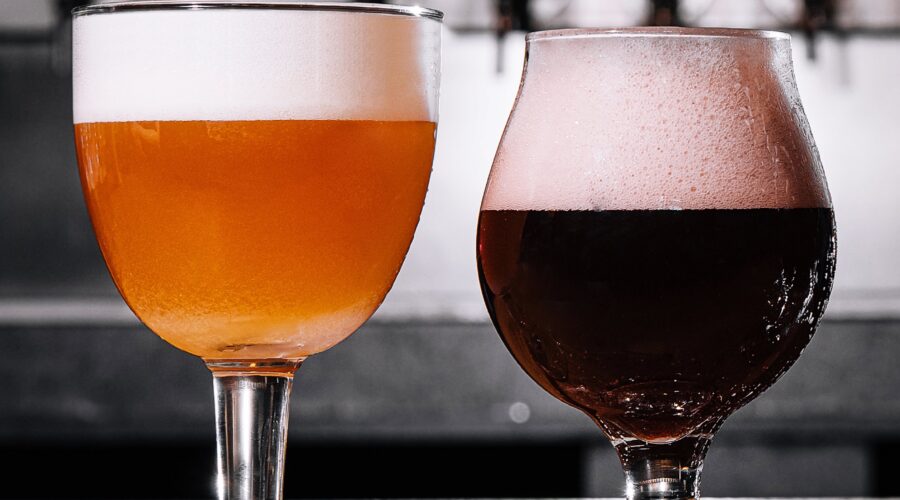 Two glasses of seasonal brews, one amber and one dark, with frothy heads, displayed on a wooden bar in front of beer taps.