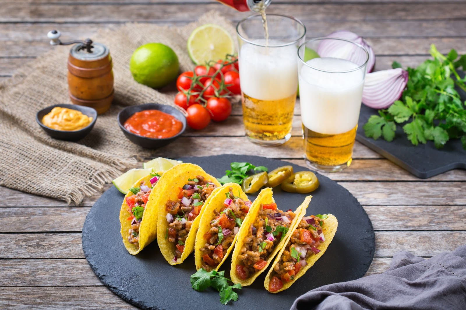  A vibrant spread of tacos filled with colorful toppings on a slate platter, accompanied by fresh ingredients, sauces, and glasses of frothy beer being poured.