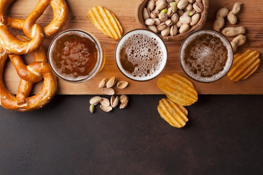 Three glasses of IPAs with pretzels, pistachios, peanuts, and chips on a wooden surface.