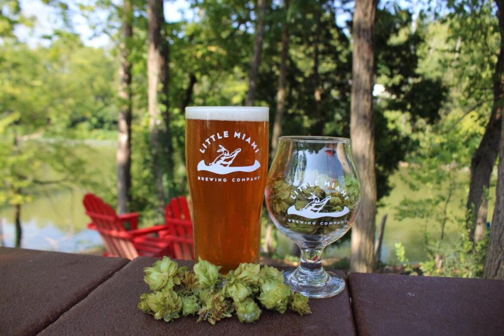 A pint glass of Little Miami Brewing Company beer with a hop flower and a tulip glass on a table overlooking a lake and trees.