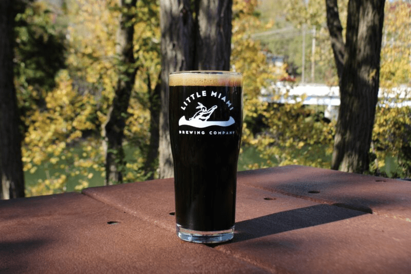A pint glass filled with dark beer sits on a wooden table outside with a scenic view of the river.