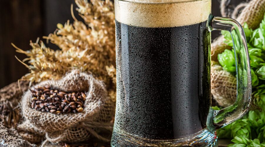 A glass of dark stout beer with barley, hops, and wheat on a rustic wooden surface.