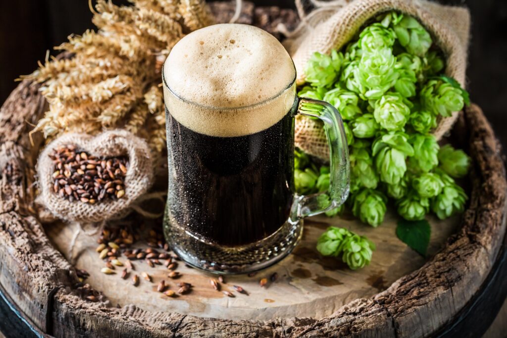 A glass of dark stout beer with barley, hops, and wheat on a rustic wooden surface.