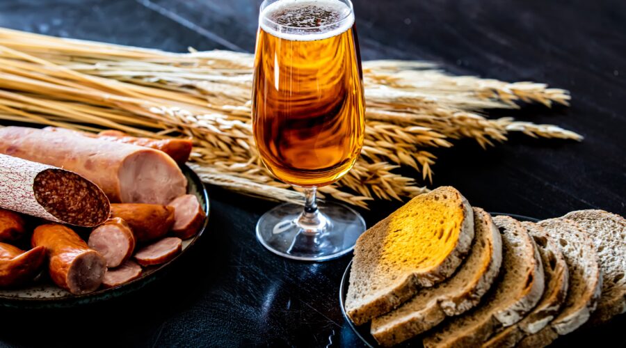 A glass of amber beer alongside sausages, bread, and wheat stalks on a dark table.