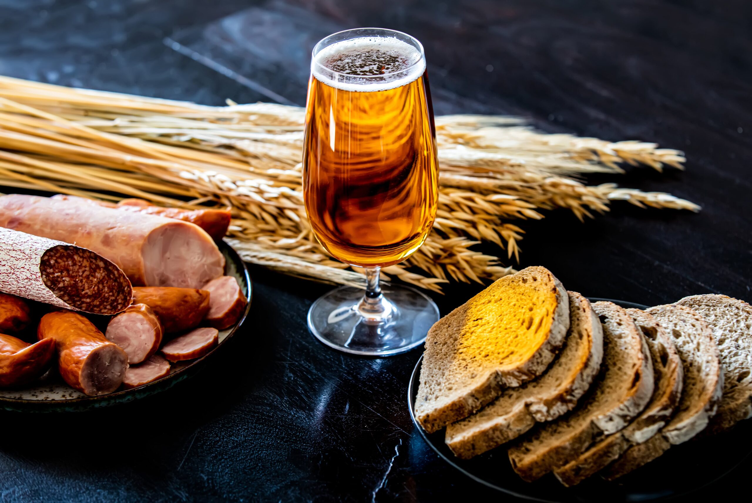 A glass of amber beer alongside sausages, bread, and wheat stalks on a dark table.