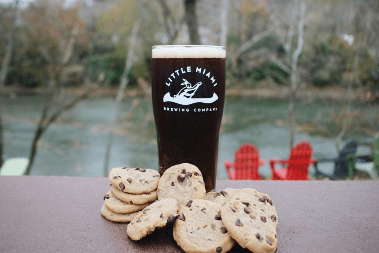 A pint glass filled with dark beer sits on a table next to a pile of chocolate chip cookies, with a scenic river view in the background.