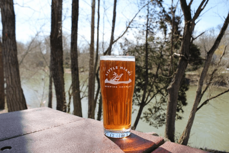 A pint glass filled with golden beer sits on a wooden table outside with a scenic view of the river.