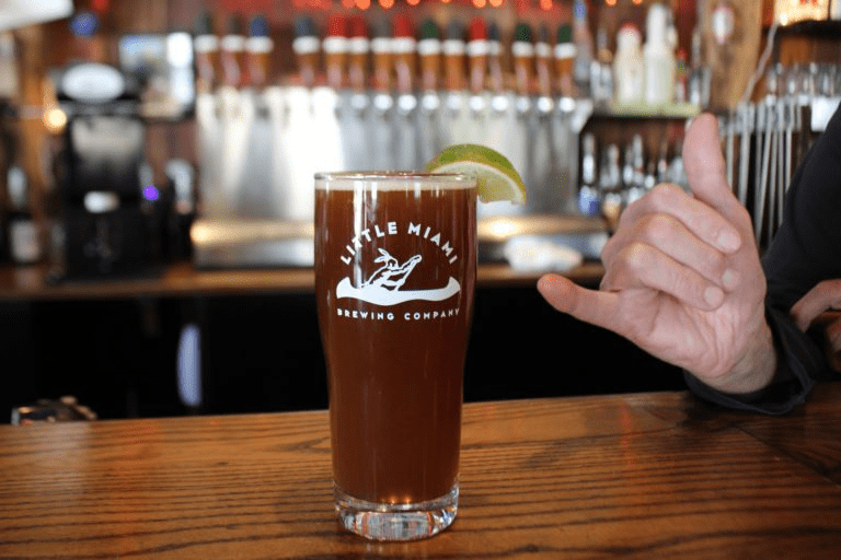 A pint glass filled with dark beer sits on a bar counter with a lime wedge and a hand making the shaka sign in the background.