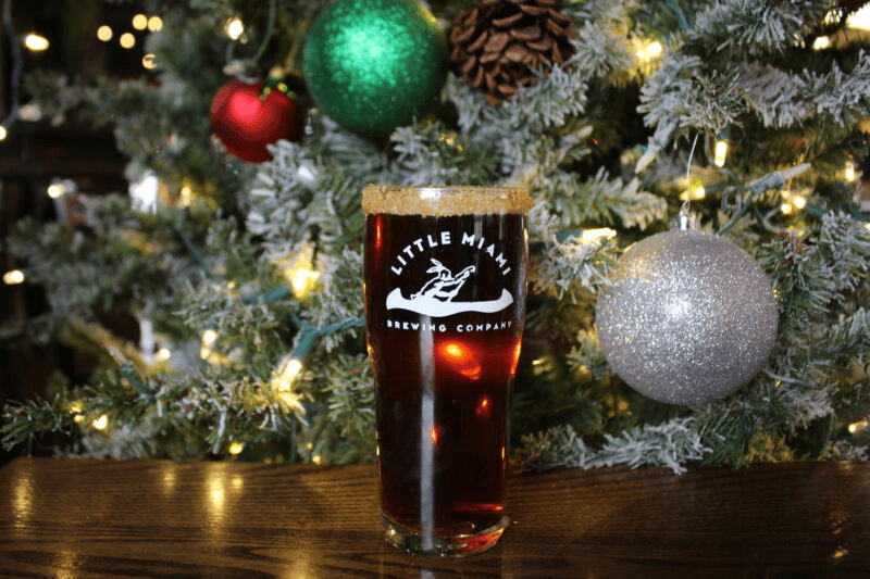 A pint glass filled with dark beer sits on a wooden table in front of a decorated Christmas tree.