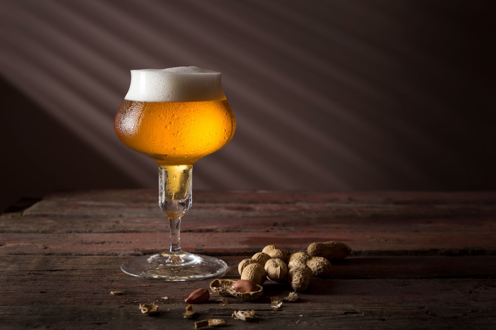 A tulip glass filled with golden Belgian beer, topped with a frothy white head, sits on a rustic wooden table alongside scattered peanuts.