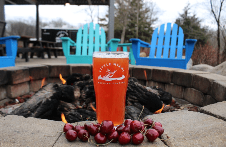 Glass of cherry-flavored craft beer by Little Miami Brewing Company placed near a cozy fire pit, perfect for seasonal enjoyment outdoors.