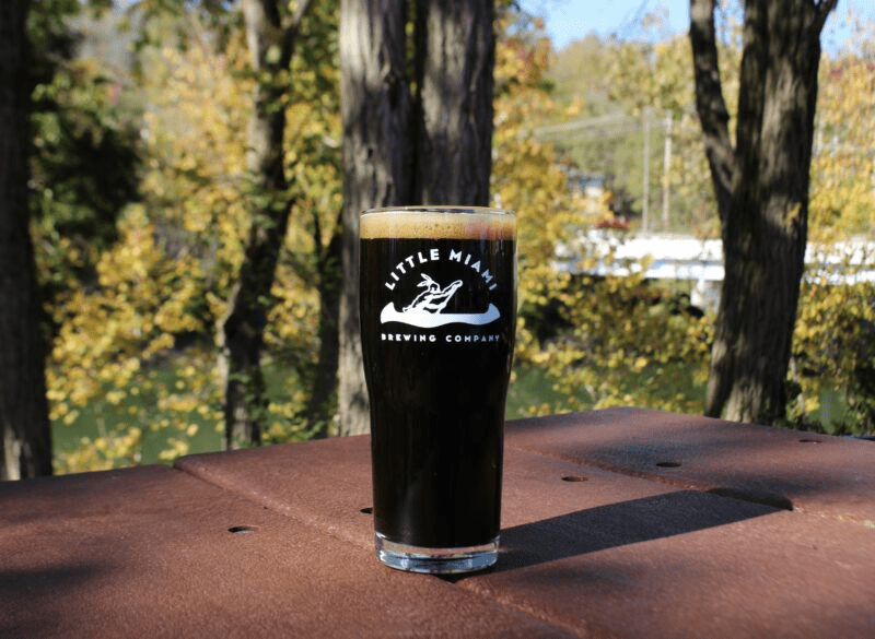 A pint glass filled with dark beer sits on a wooden table outside with a scenic view of the river.

