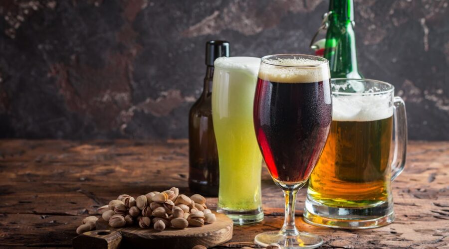 A variety of craft beers served in different glassware, accompanied by a bowl of pistachios on a rustic wooden table.