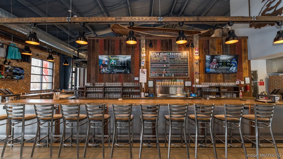 A cozy brewery bar with a rustic wooden design featuring a long wooden countertop lined with high metal chairs. Behind the bar, a chalkboard displays the beer menu from "Little Miami Brewing Co." with taps below it. Two mounted TVs on either side of the chalkboard display sports games. The space is warmly lit with industrial-style pendant lights, and the exposed brick walls and ductwork add to the brewery's inviting and industrial-chic aesthetic.
