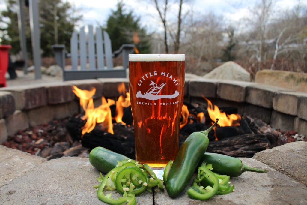 A close-up shot of a pint glass of beer from Little Miami Brewing Company. The glass is placed on a stone ledge in front of a cozy outdoor fireplace. The amber beer has a frothy head, and beside the glass, there are sliced jalapeno peppers, suggesting that the beer flavor is spicy. 
