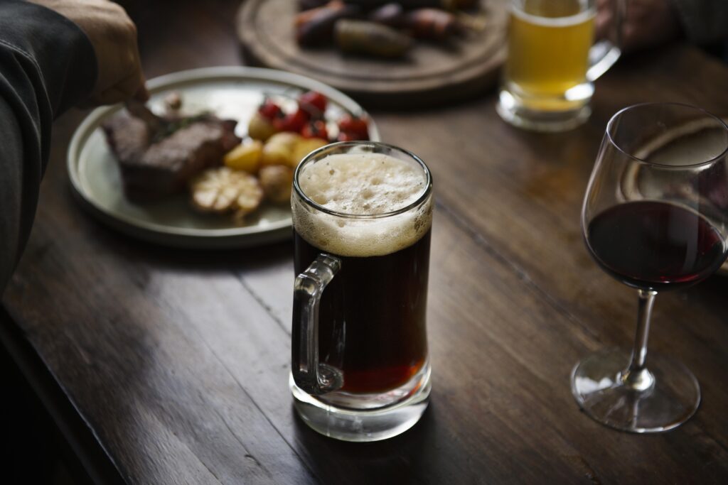 A glass of beer on a table with a plate of food and a glass of red wine.