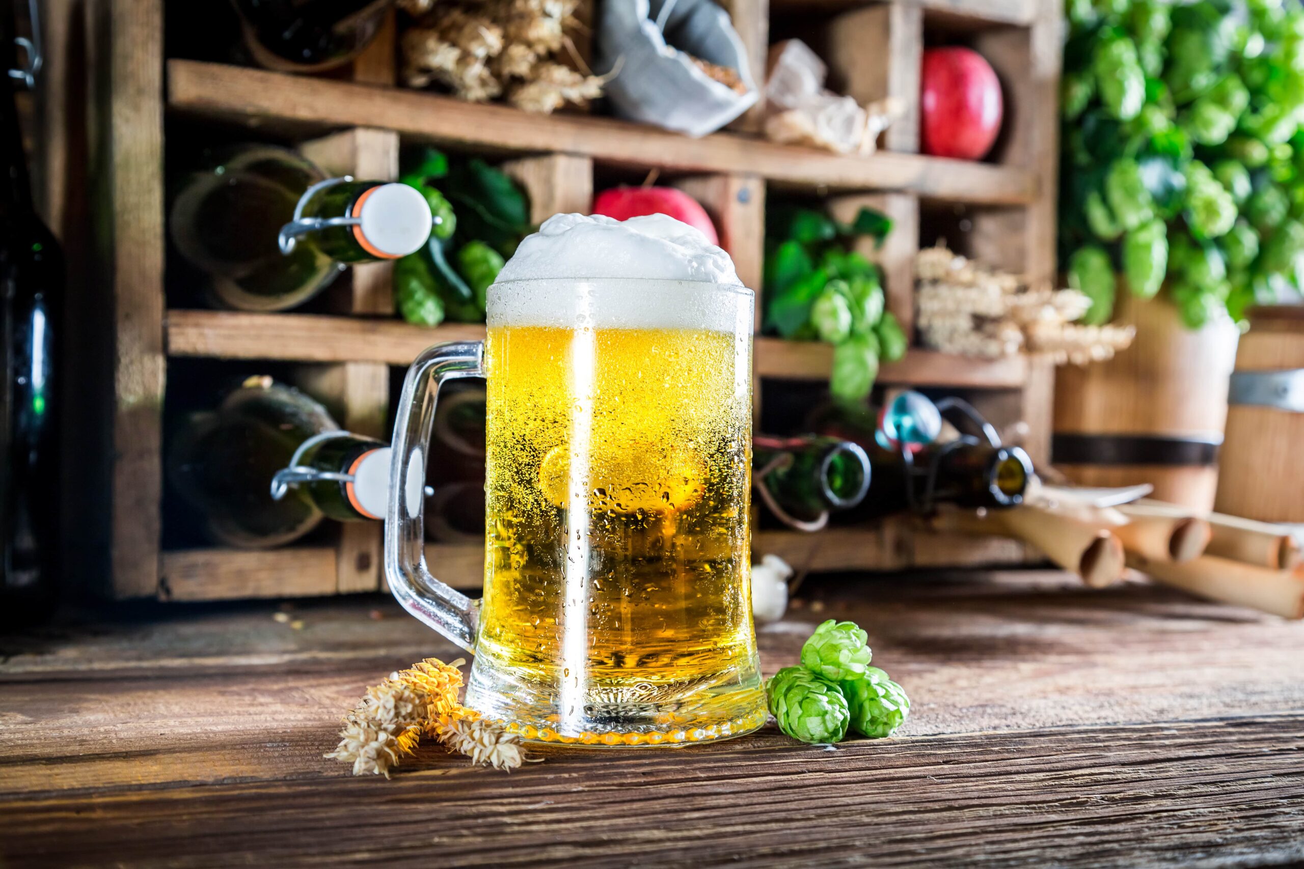 A glass of beer with a thick head of foam, surrounded by hops and other brewing ingredients.