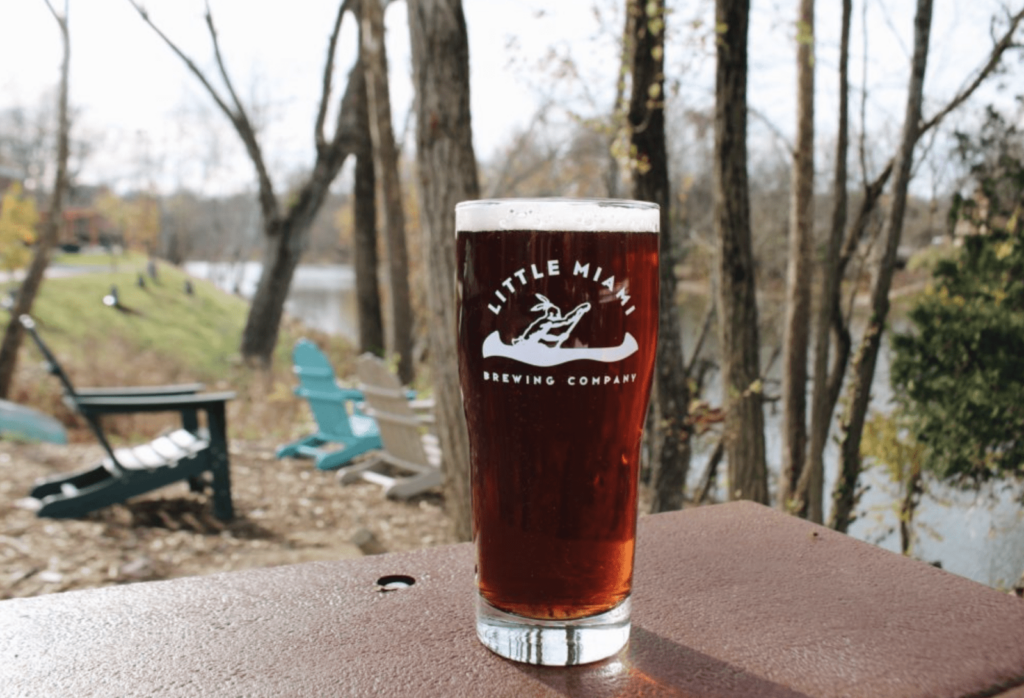 A glass of Little Miami Brewing Company beer on a table outdoors, with trees and a river in the background.