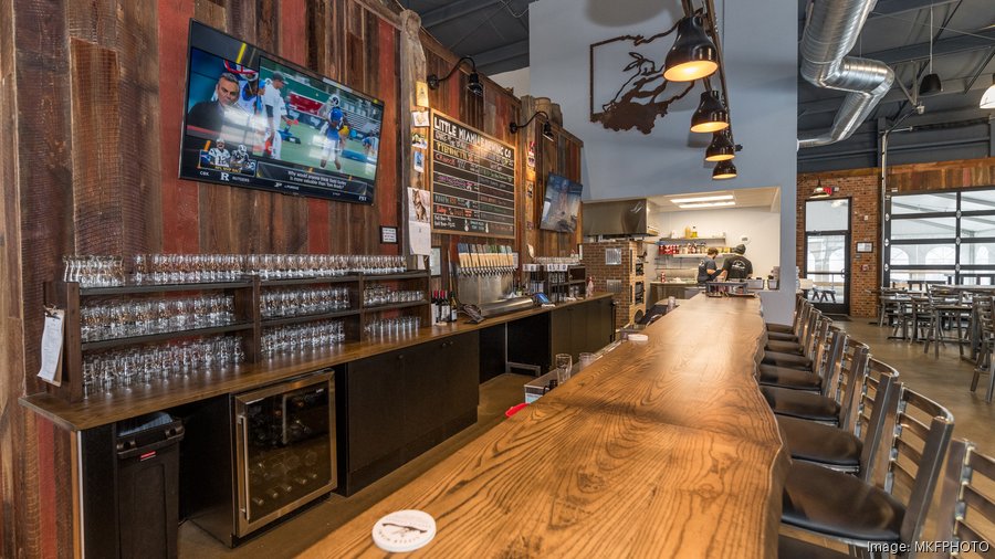 This image captures the inviting atmosphere of a pub. A tall glass of beer with a foamy head rests on a wooden bar counter, illuminated by warm lighting. The background is blurred, suggesting a lively and bustling environment.