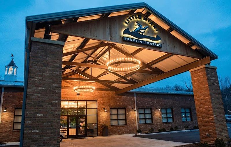 The exterior of Little Miami Brewing Company during the evening, showcasing a welcoming entrance with a timber-framed overhang and warm glowing circular chandeliers.