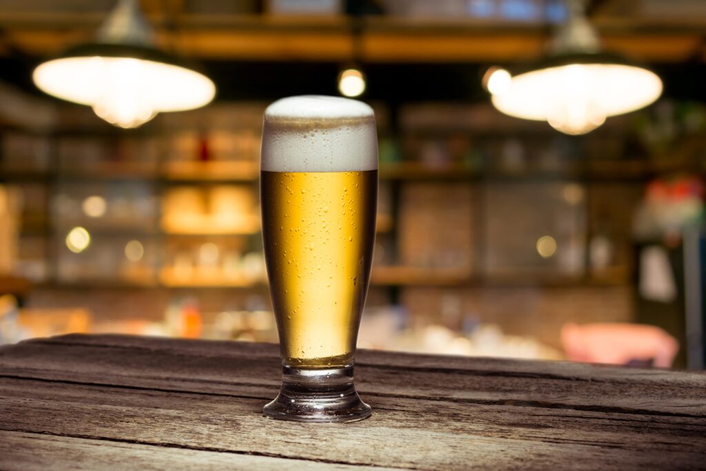 A tall glass of beer with a thick head of foam, sitting on a wooden bar counter.